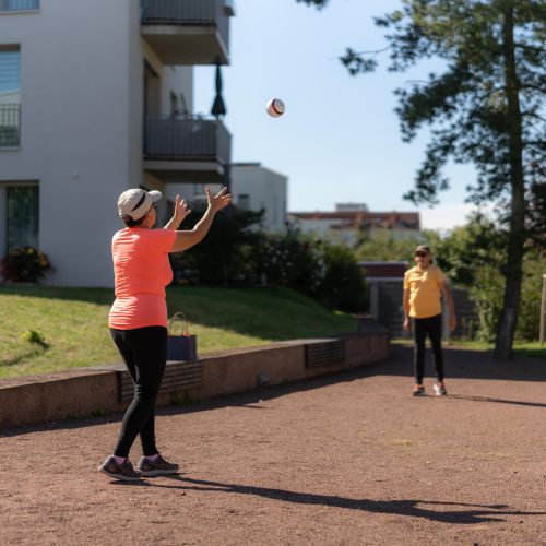 Zwei Menschen spielen draußen an einem sonnigen Tag in der Nähe eines Wohnhauses Fangen mit einem Ball.